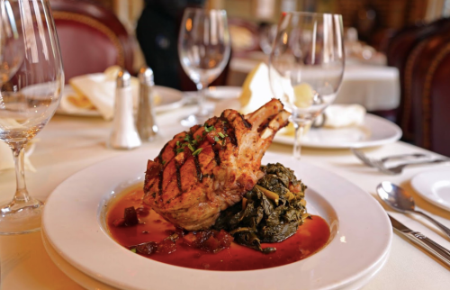 A beautifully plated pork chop with greens and sauce, set on a fine dining table with elegant glassware.