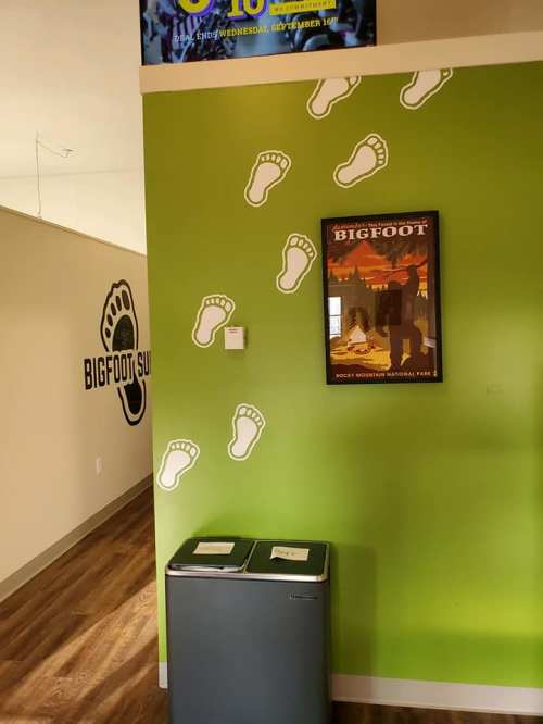 A green wall with Bigfoot footprints leading to a poster of Bigfoot in a room with a trash bin.