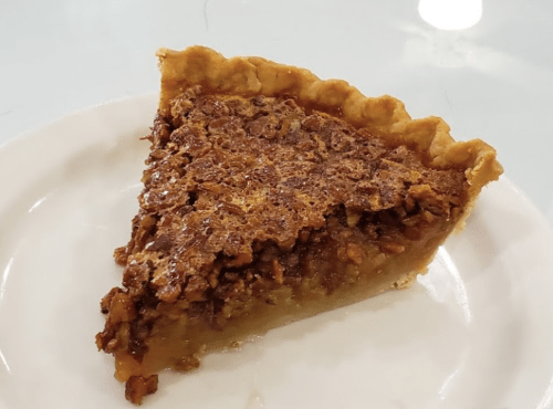 A slice of pecan pie on a white plate, featuring a golden crust and a rich, nutty filling.