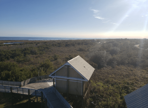 A coastal view from above, featuring a wooden structure, lush greenery, and the ocean under a clear blue sky.
