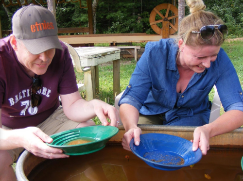 Two people pan for gold in a shallow tub, smiling and focused on their activity outdoors.