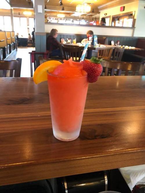 A frozen drink in a glass, garnished with an orange slice and a strawberry, sits on a wooden table in a restaurant.