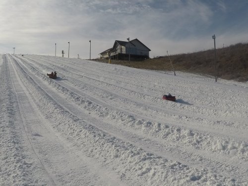 Two people sledding down a snowy hill with a lodge in the background under a cloudy sky.