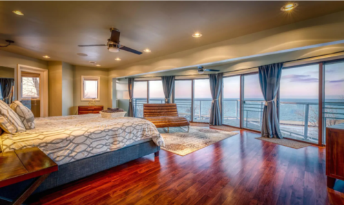 Spacious bedroom with large windows overlooking a lake, featuring a bed, wooden furniture, and modern decor.