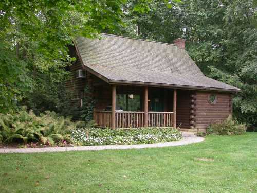 A cozy log cabin surrounded by greenery and flowers, featuring a porch and a sloped roof.