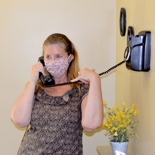 A woman wearing a mask talks on a vintage telephone in a room with yellow flowers and a wall clock.