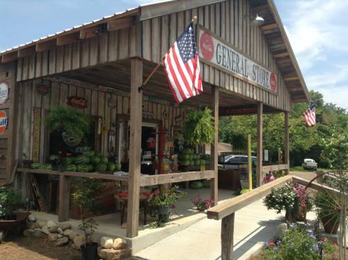 A rustic general store with American flags, watermelons, and colorful flowers on a sunny day.