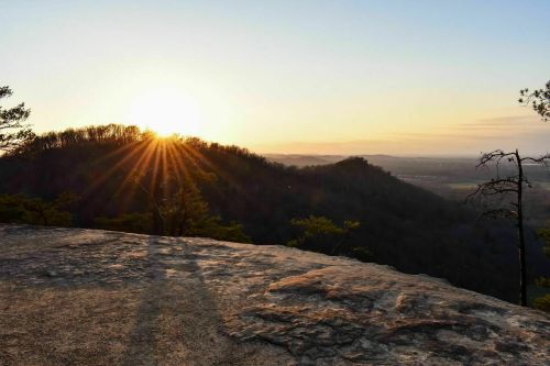 Sunrise over a mountainous landscape, casting rays of light and shadows on a rocky outcrop.