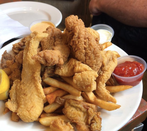 A plate piled high with fried fish, chicken, and golden fries, accompanied by dipping sauces and a lemon wedge.