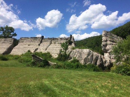 A rocky cliff formation with lush green grass and trees under a blue sky with fluffy white clouds.