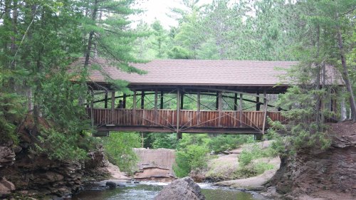 A wooden covered bridge spans a creek, surrounded by lush green trees and rocky terrain.