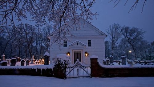 A snowy cemetery at dusk, featuring a white building and a brick gate surrounded by snow-covered trees and gravestones.