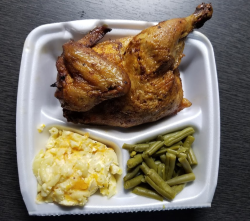 A takeout container with a roasted chicken, creamy mashed potatoes, and green beans on a dark wooden surface.