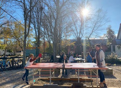 Children playing ping pong at a sunny outdoor table, with people socializing in the background among trees.