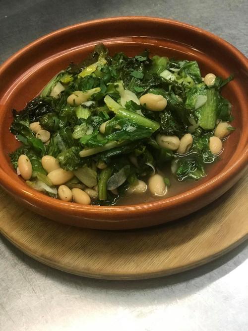 A bowl of mixed greens and white beans in a light broth, served on a wooden platter.