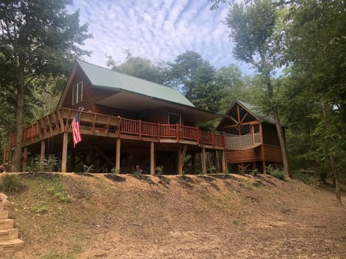 A wooden cabin with a green roof, elevated on stilts, surrounded by trees and featuring a porch and an American flag.