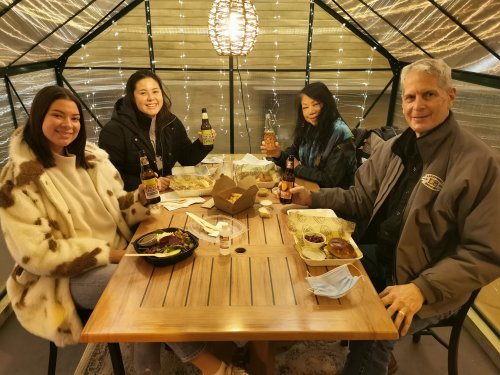 Four people sitting at a wooden table in a cozy setting, enjoying food and drinks, with warm lighting around them.