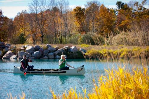 Two people paddle a canoe on a calm lake surrounded by autumn foliage and rocky shores.