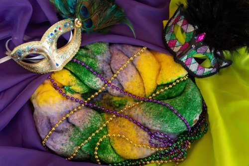 A colorful King Cake surrounded by Mardi Gras masks and beads on vibrant fabric.