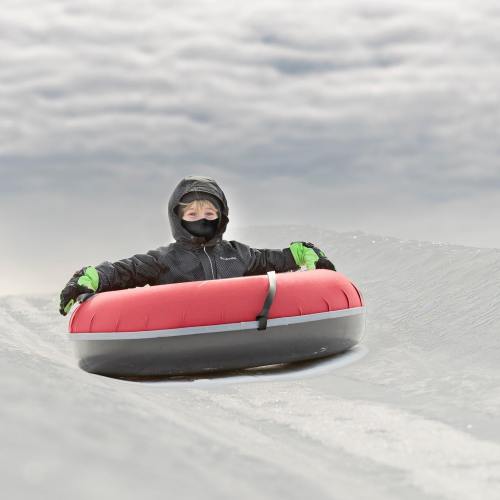 A child in a black jacket and mask sits in a red snow tube, ready to slide down a snowy hill.