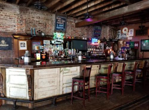 A rustic bar with a wooden counter, brick walls, and various bottles displayed on shelves, featuring bar stools and neon signs.