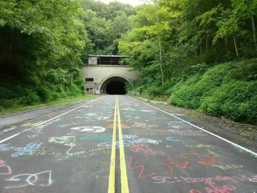 A colorful, graffiti-covered road leads to a dark tunnel, surrounded by lush green trees.