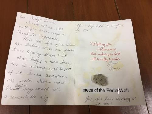 A handwritten letter with a Christmas greeting and a piece of the Berlin Wall attached, resting on a wooden surface.