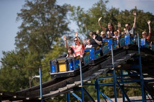 A group of people on a roller coaster, raising their hands in excitement as they ride through a sunny amusement park.