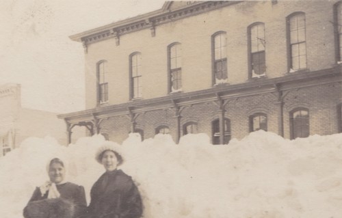 Two women stand in front of a large snowbank, with a brick building in the background, in a vintage black-and-white photo.