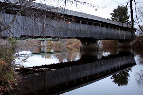 A wooden covered bridge spans a calm river, reflecting its image in the water, surrounded by autumn foliage.
