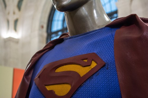 Close-up of a superhero costume featuring a blue shirt with a red cape and a yellow and red "S" emblem.