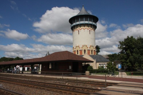 Historic train station with a tall, round water tower, surrounded by trees and a blue sky with fluffy clouds.
