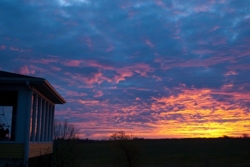 A vibrant sunset with pink and orange hues fills the sky, viewed from the side of a house against a grassy field.