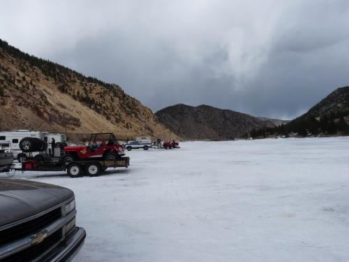 A snowy landscape with parked vehicles and trailers near a frozen lake, surrounded by mountains and cloudy skies.