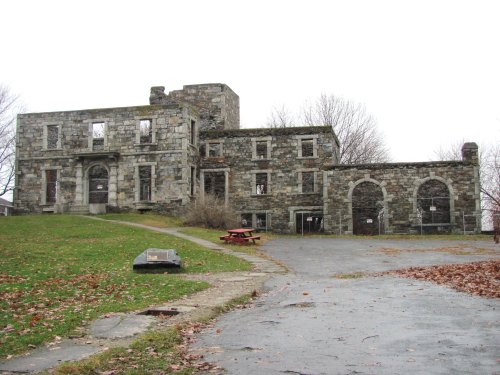 A weathered stone building with multiple windows and a grassy area, surrounded by bare trees and fallen leaves.