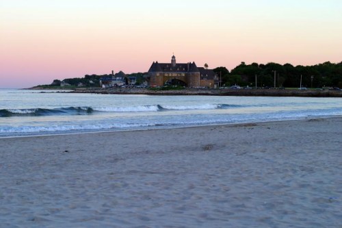 A serene beach at sunset, with gentle waves and a historic building in the background against a pastel sky.