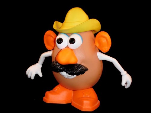 A toy Mr. Potato Head with a yellow cowboy hat, orange shoes, and a black mustache against a black background.