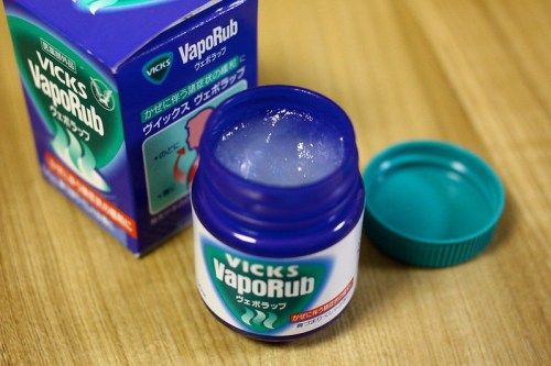 A jar of Vicks VapoRub with the lid off, next to its box, on a wooden surface.