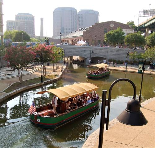 Two boats with passengers navigate a scenic canal, surrounded by greenery and urban buildings in the background.