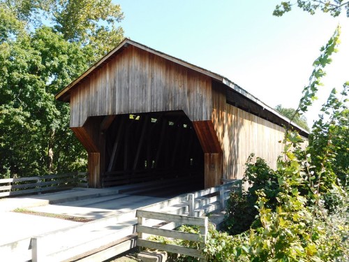 A wooden covered bridge surrounded by trees, with a clear blue sky above and a gravel path leading to it.