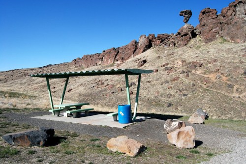 A picnic area with a green shelter and blue trash can, set against rocky hills under a clear blue sky.