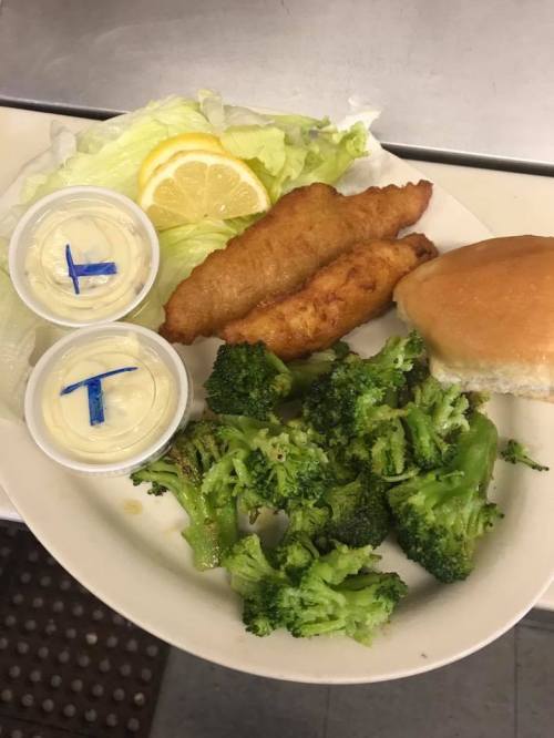 A plate with two fried fish fillets, broccoli, a bun, a lemon slice, and two cups of sauce.