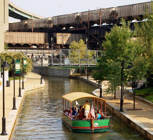 A boat with passengers glides through a canal beneath a railway bridge, surrounded by greenery and urban structures.