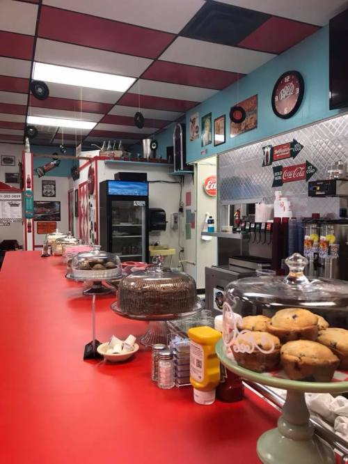 A retro diner interior with a red counter, glass display cases, and vintage decor, featuring baked goods and condiments.