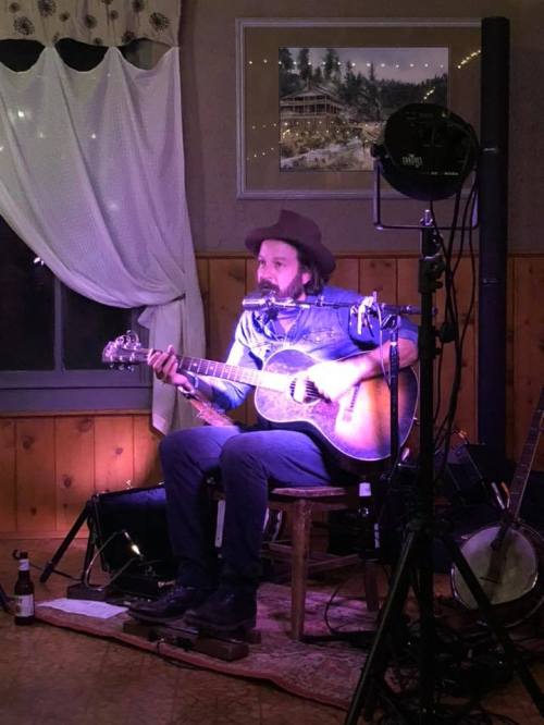 A musician in a hat plays guitar on stage, seated on a chair with a microphone and warm lighting in a cozy venue.