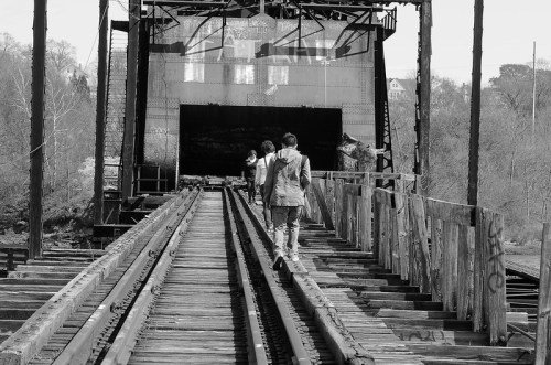 Two people walk along an old, wooden railway bridge leading to a dark tunnel, surrounded by trees.