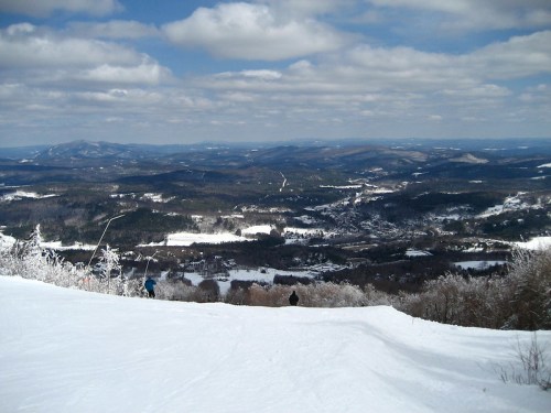 A snowy landscape with rolling hills and a valley under a partly cloudy sky, viewed from a mountain slope.