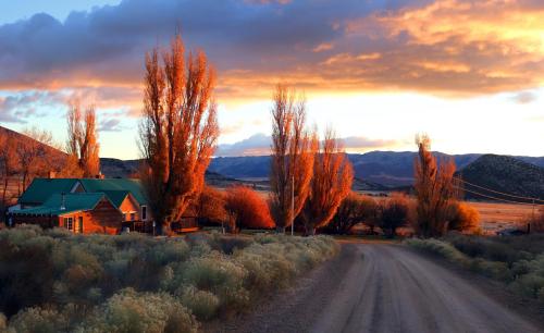 A dirt road leads to a rustic house surrounded by colorful autumn trees and a scenic mountain backdrop at sunset.