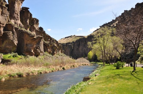 A serene river flows through a canyon, surrounded by lush greenery and rocky cliffs under a clear blue sky.