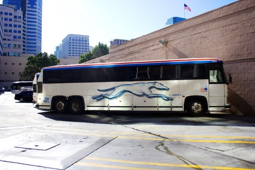 A Greyhound bus parked beside a building in an urban area, with a clear blue sky above.